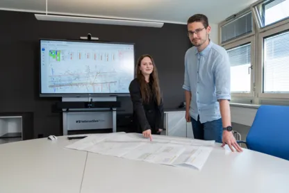 Zwei Personen stehen lächelnd an einem Tisch, eine Person zeigt auf einen Plan auf dem Tisch.