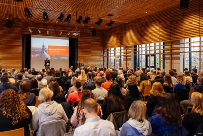 Eine Momentaufnahme vom Personalforum 2024. Personen sitzen mit dem Rücken zugewandt und hören einem Vortrag des Personalforums zu. Im Hintergrund ist eine Tribüne mit einer Präsentation.