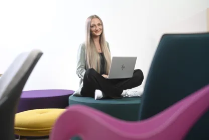 Eine Person sitzt auf einem Hocker und lächelt in die Kamera, auf dem Schoß hat sie ein Laptop.
