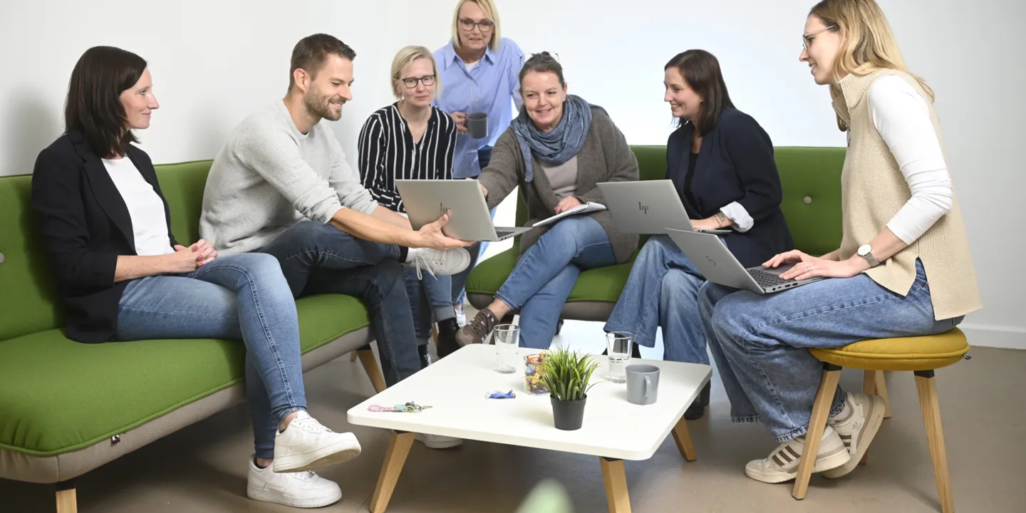 Eine Gruppe Menschen sitzt auf Sofas und schaut sich etwas auf dem Laptop an.